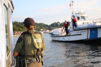 A conservation officer stands on a boat, looking toward another law enforcement vessel where two people wearing orange lifejackets stand ready.