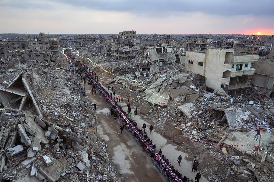 People gather around a long, narrow table surrounded by the rubble of destroyed homes and buildings.