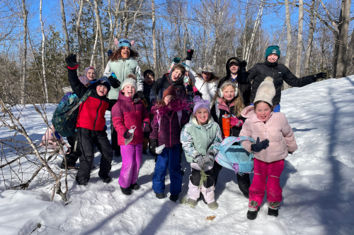 A crowd of more than 10 children in winter coats and snowpants and smiling and waving from a snowy wooded hill.