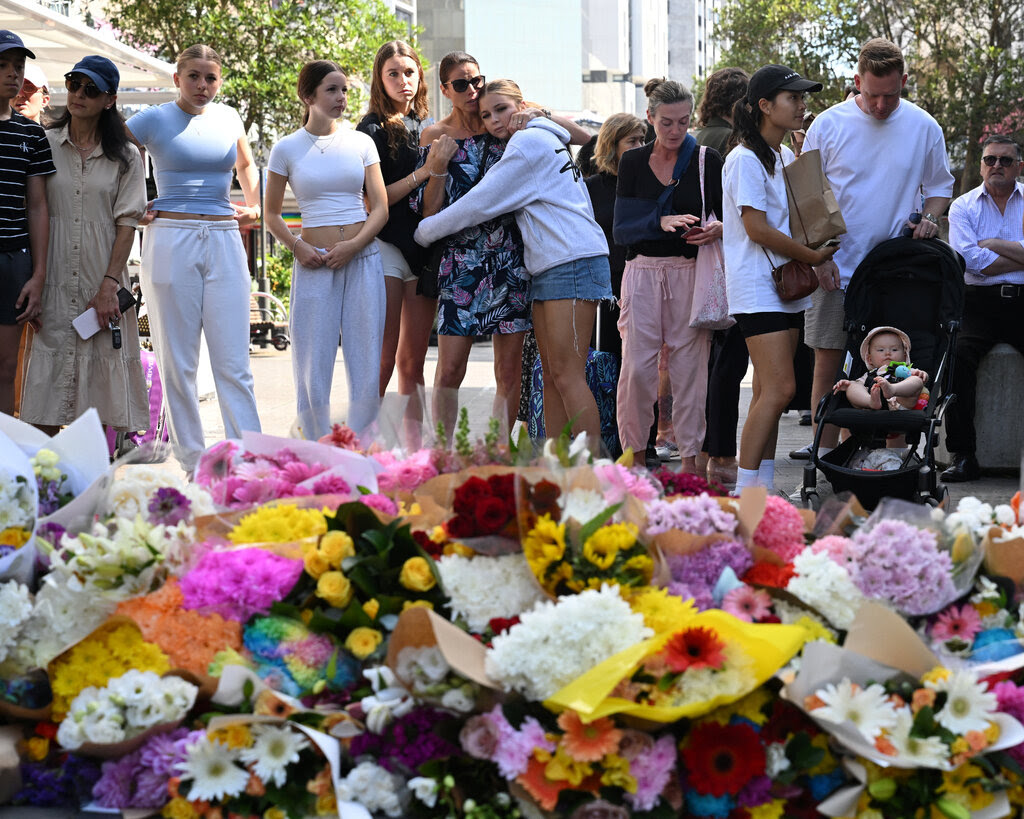 At least a dozen people mourning in front a big pile of flower bouquets. 