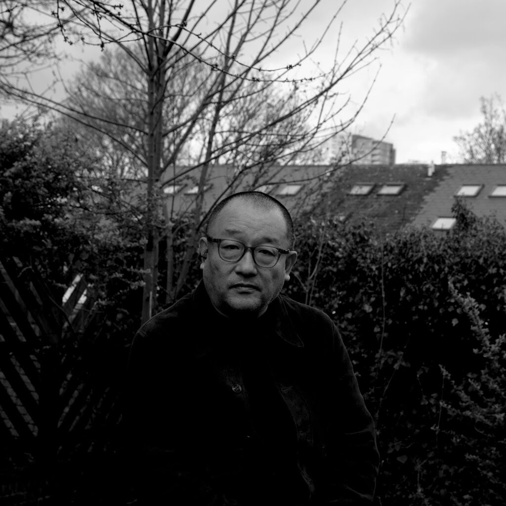 Wang Xiaoshuai, wearing black clothes and sitting on a stool outdoors in front of a spindly tree without leaves on a gray day with an overcast sky. 