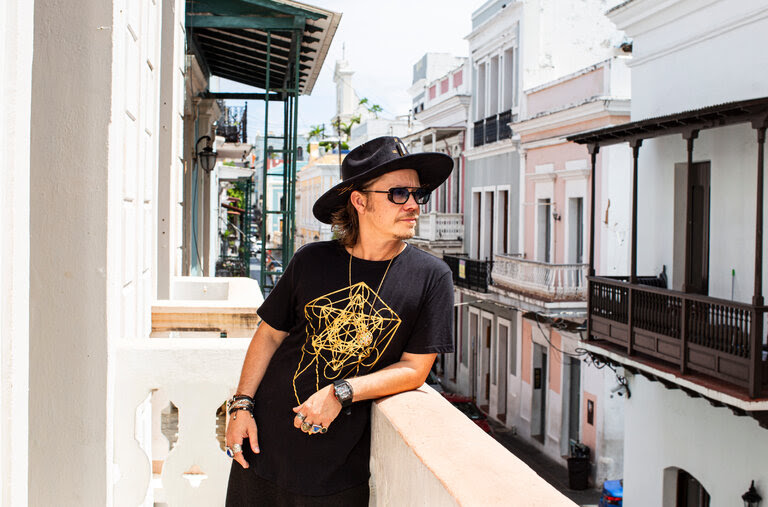 Brock Pierce, wearing a black T-shirt, black hat and lots of bracelets and rings, stands on a balcony on a city street.