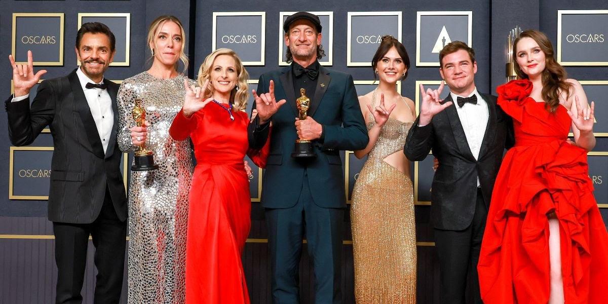 Seven people, comprising the key cast members and writer-director of the film CODA, two of whom hold Academy Awards, stand in front of a step and repeat backdrop doing the I Love You sign in American Sign Language.
