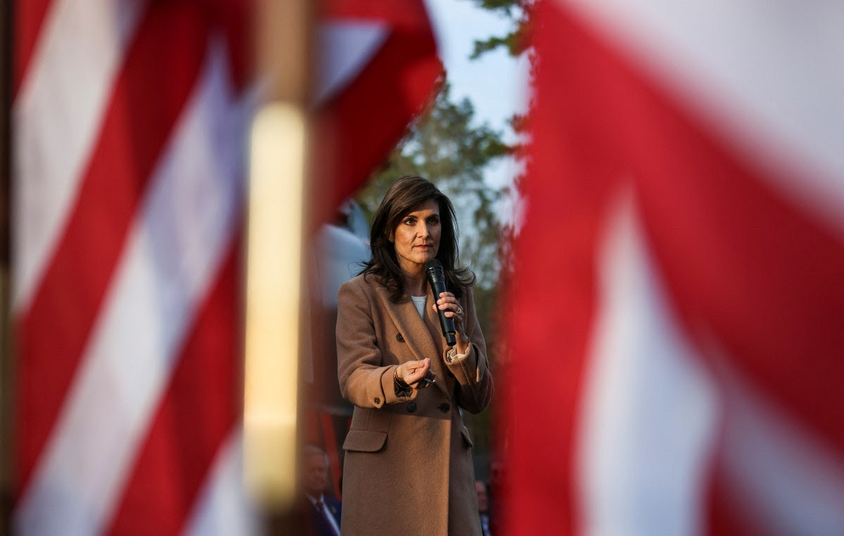 Republican presidential candidate Nikki Haley photographed in between two American flags. Haley wears a brown coat, holds a microphone in her left hand, and is gesturing with her right hand.
