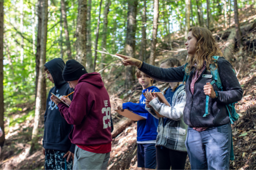 A white woman points at a tree during an outdoor lesson with teens.