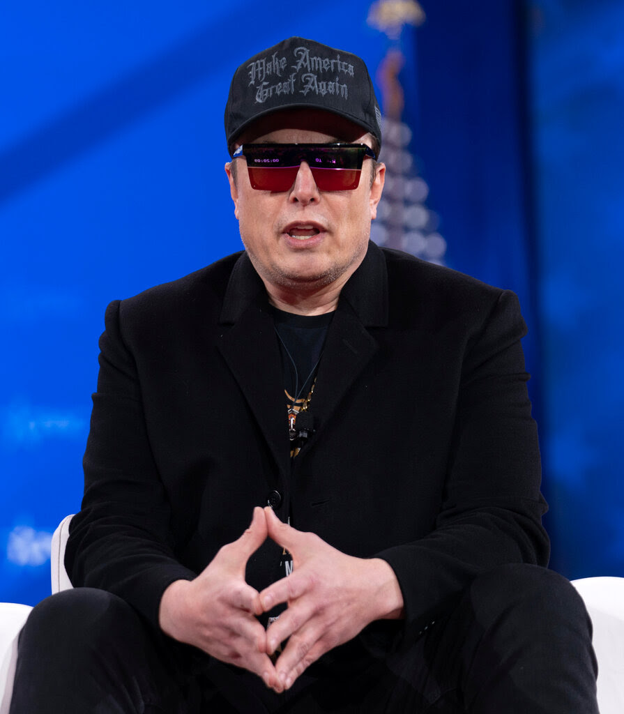 Elon Musk in a black MAGA hat, a black blazer and sunglasses.