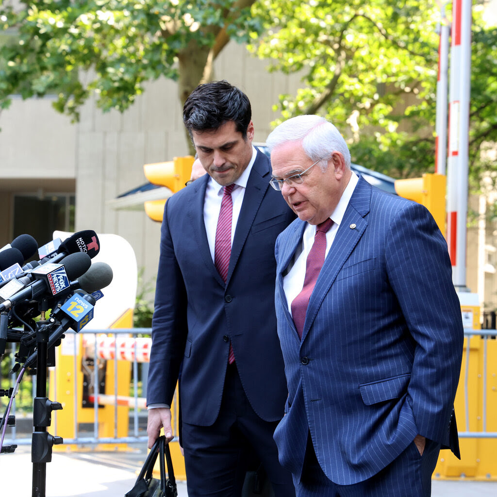 Senator Robert Menendez, in a blue suit and red tie, leaves Federal District Court in Manhattan. In front of him are microphones from news media. 