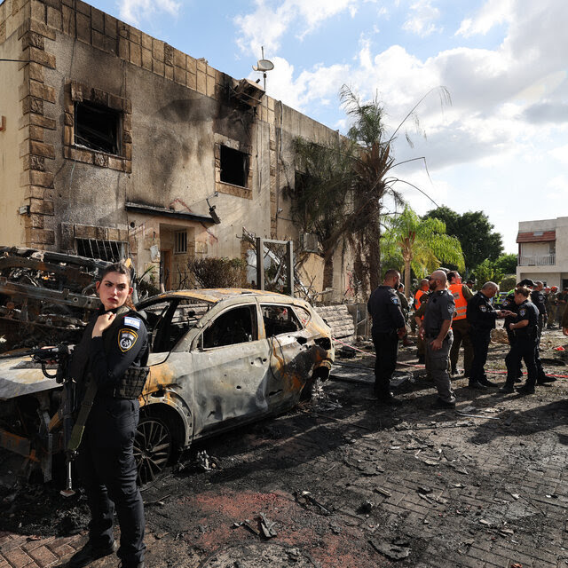 A woman in uniform standing near a charred car and a damaged building. Other officials, some in orange vests, are nearby.