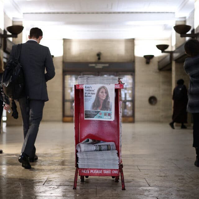 Men and women dressed in dark colors walk past a bright red newsstand with stacked copies of The Evening Standard, whose front page features a photo of Britain’s Catherine, Princess of Wales, at a subway station.