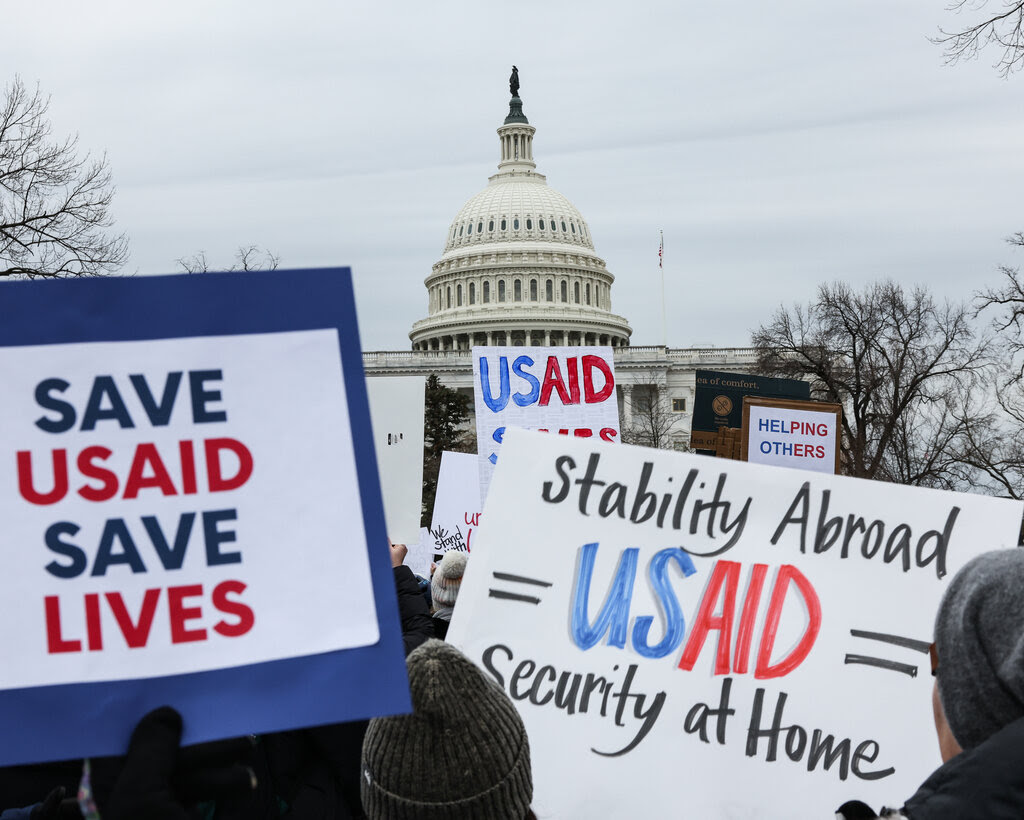 Protest signs outside the Supreme Court read: “Save U.S.A.I.D. Save Lives” and “Stability Abroad. U.S.A.I.D. Security at home.” Bare tree branches frame the protesters.