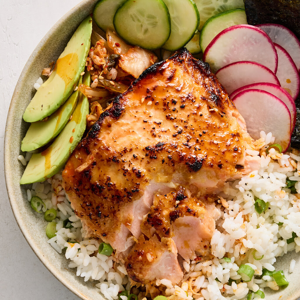 Broiled salmon on a bowl of steamed rice with scallions and slices of cucumber, radish and avocado.