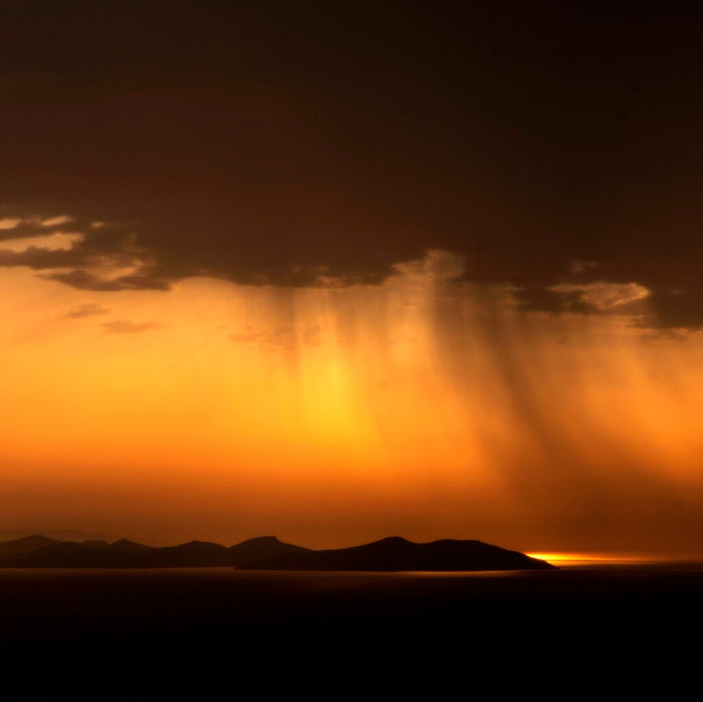 An island at sunset is depicted as a dark, hilly silhouette beneath a sky that is various shades of orange. Above the orange section of the sky are dark clouds. The sea is also dark, except for directly around the island, where the sea appears orange from reflected light.