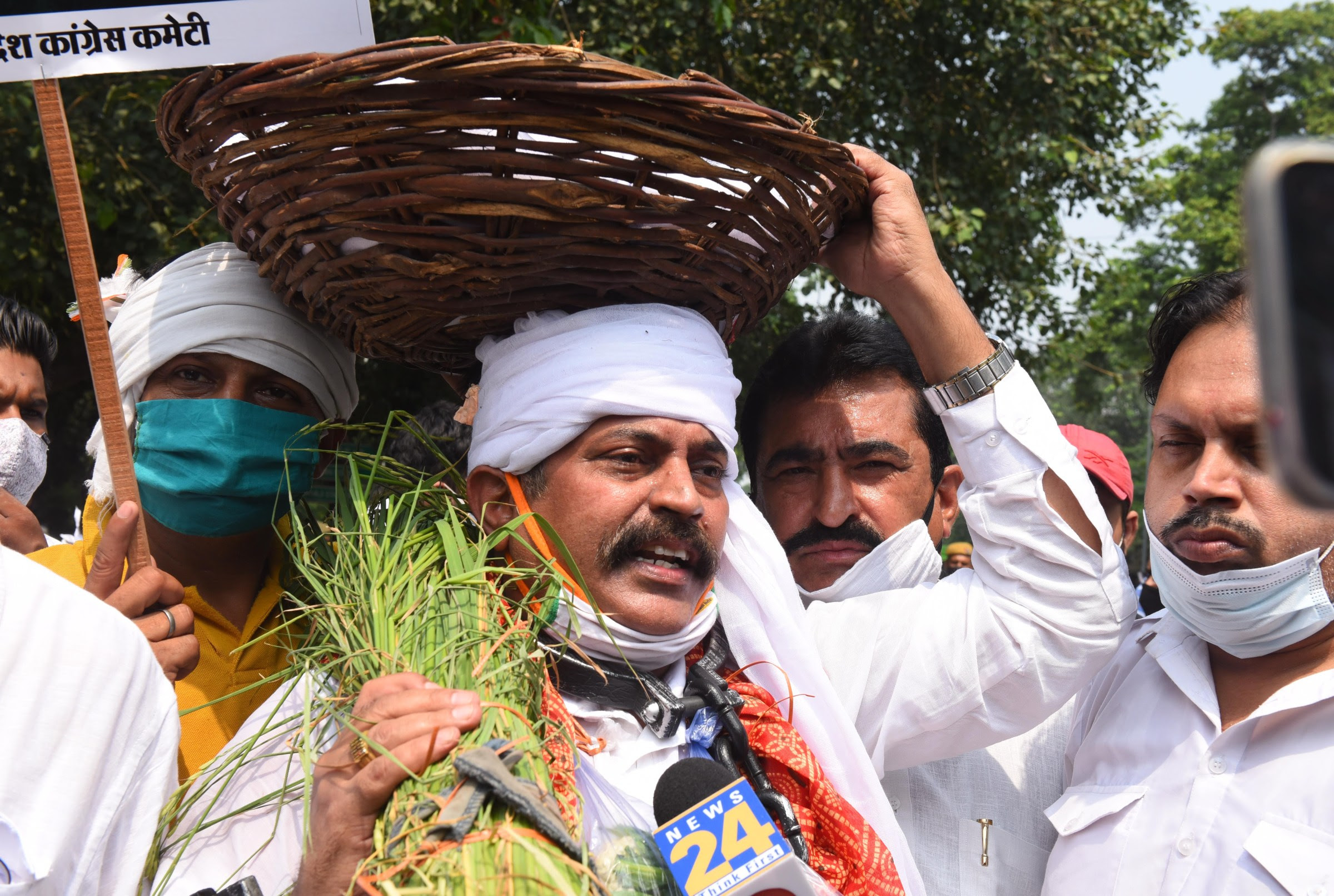 An Indian man carrying a basket on his head and a sheaf of grass in one hand stands in a crowd and speaks into a news microphone.