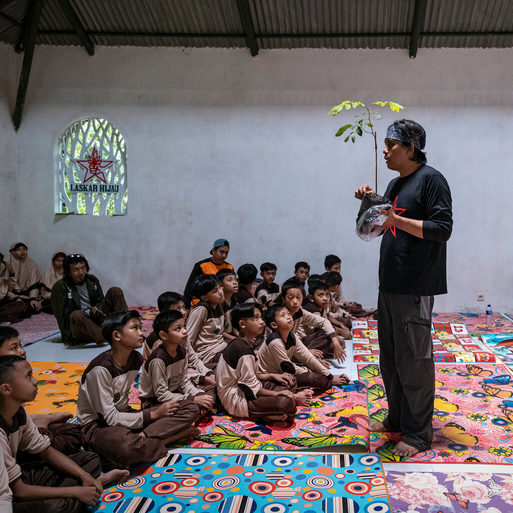 A man holding a sapling in his hands and talking a group of children sitting on vividly colored blankets inside a building. 