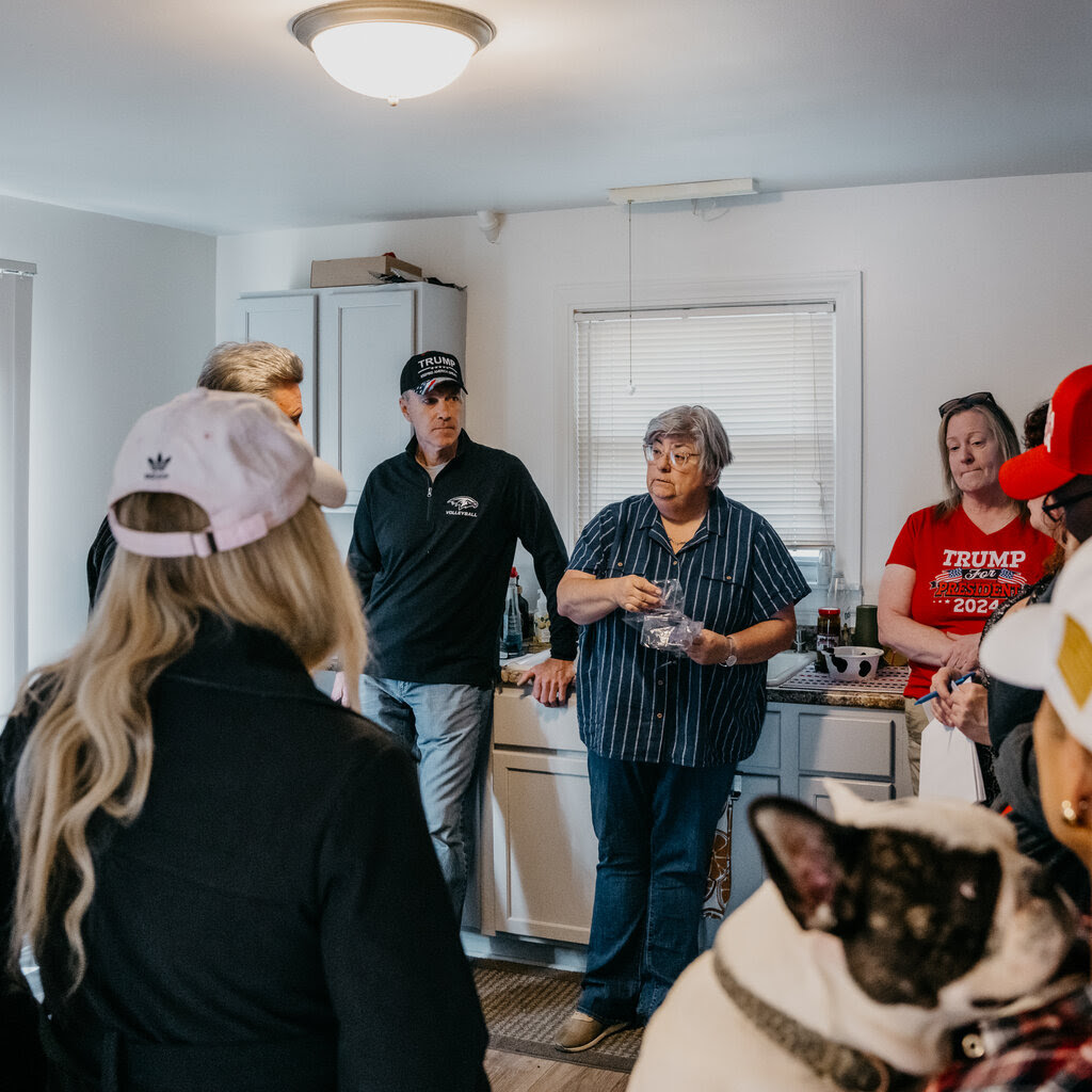 Several people stand around in a kitchen. One wears a Trump campaign shirt. 