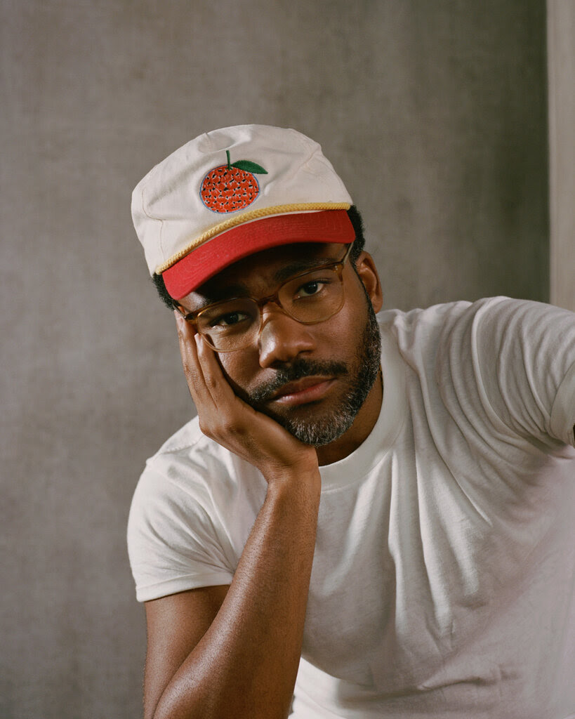 Donald Glover wearing brown glasses and a ball cap with a picture of an orange on it, rests his head on one hand and looks at the camera.