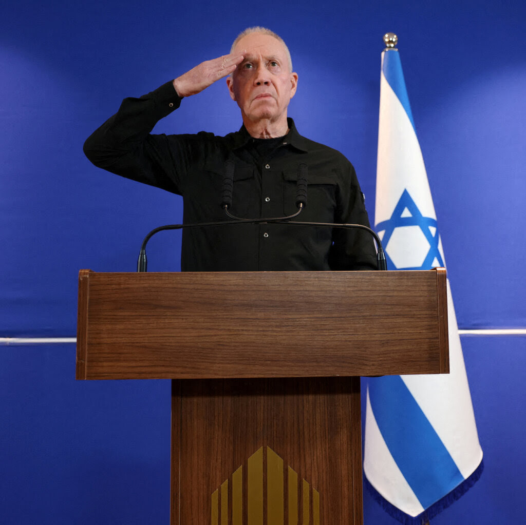 Yoav Gallant stands behind a lectern and salutes.