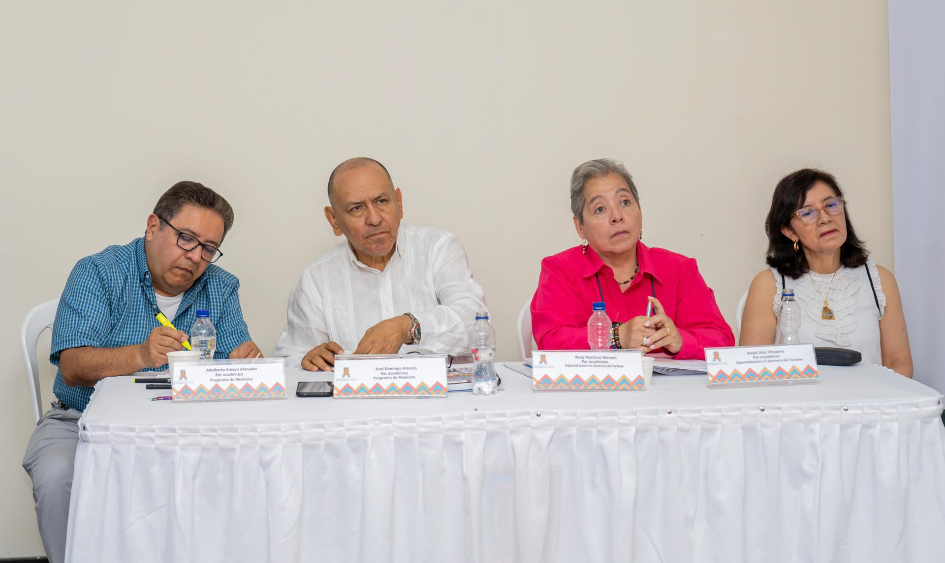 una mesa con cuatro personas sentadas, dos hombres y dos mujeres, frente a micrófonos y rótulos de identificación. Parecen estar en una mesa de panel o conferencia. Visten de manera formal y tienen expresiones serias mientras observan o toman notas.