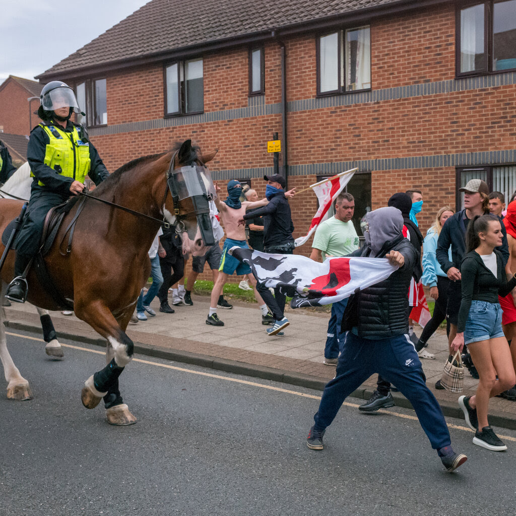 A group of people, some waving England flags and others in balaclavas, hemmed on to a pavement by mounted police.