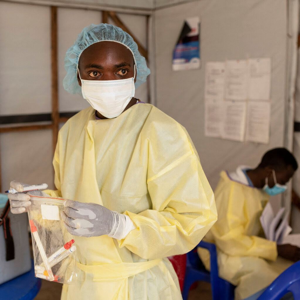 A nurse waring a yellow gown, a blue hairnet and a white face mask, holding a plastic bag with vials.