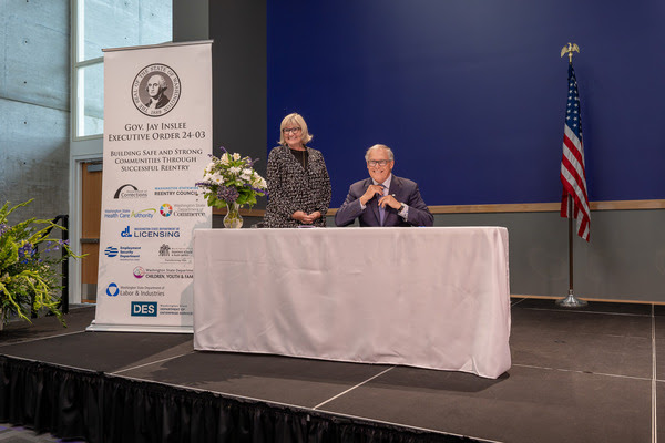 Gov Inslee and First Spouse Trudi Inslee in front of a table and banner