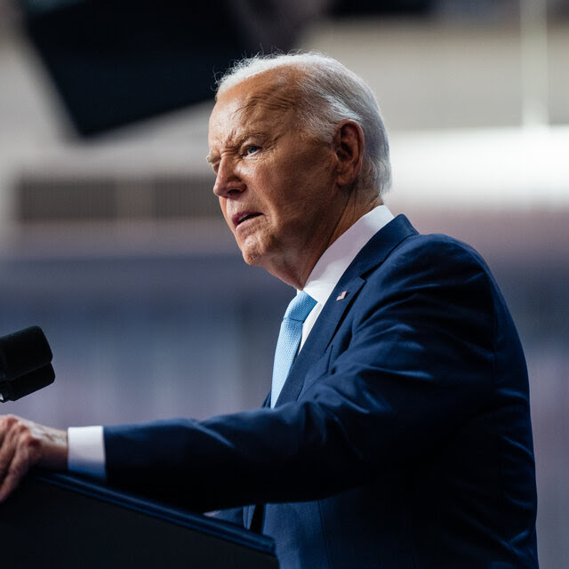 A profile view of President Biden speaking at a podium while wearing a blue suit.