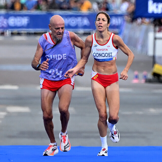 A man wearing a bib that says “Guide” running with a woman on a marathon course. 