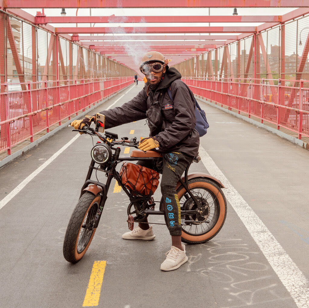 Eric Edwards, wearing goggles and a leather jacket, stands on a bridge with his homemade electric bike.
