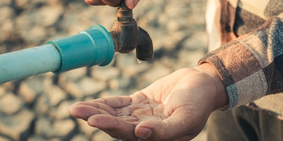 A person standing on dry, drought-stricken land holds their hand under a tap catching droplets of water.