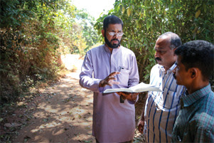 Three men are standing on a shaded pathway. One of the men has his Bible open and is speaking with the other two.