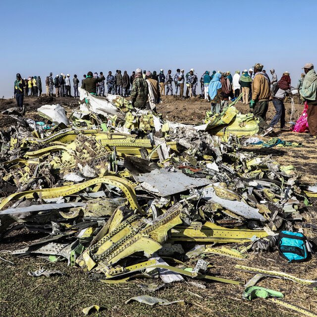 Wreckage of the 737 Max plane crash in Ethiopia is visible in the foreground and a crowd of people is visible in the background.