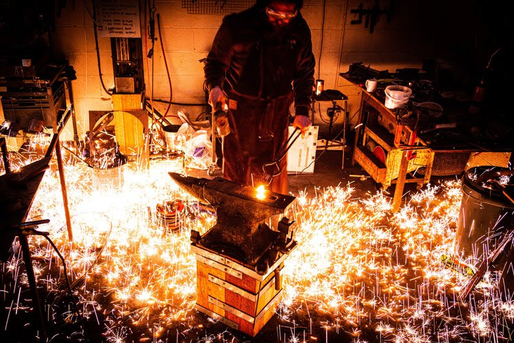 A blacksmith from the St Clement's Festival working in their forge. There are embers and sparks surrounding the anvil.