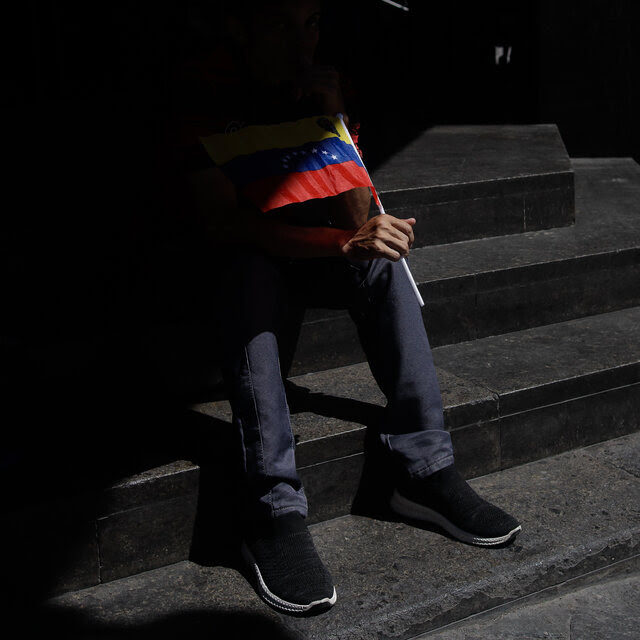 A man in shadow, holding a Venezuelan flag. All that can be seen are his legs, his feet, a corner of the flag, and a section of the black steps on which he is sitting. 
