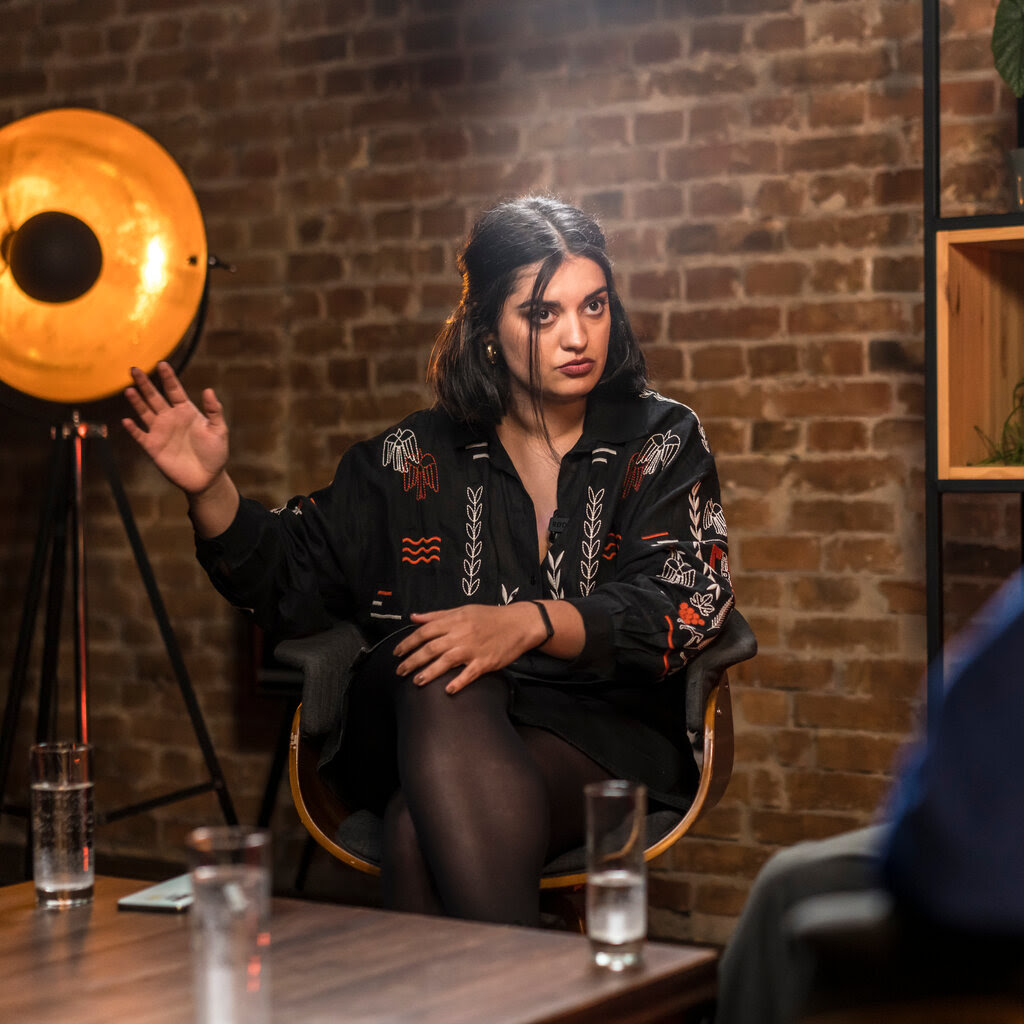 A person sitting on a chair talking to another person, who is partially in the frame.