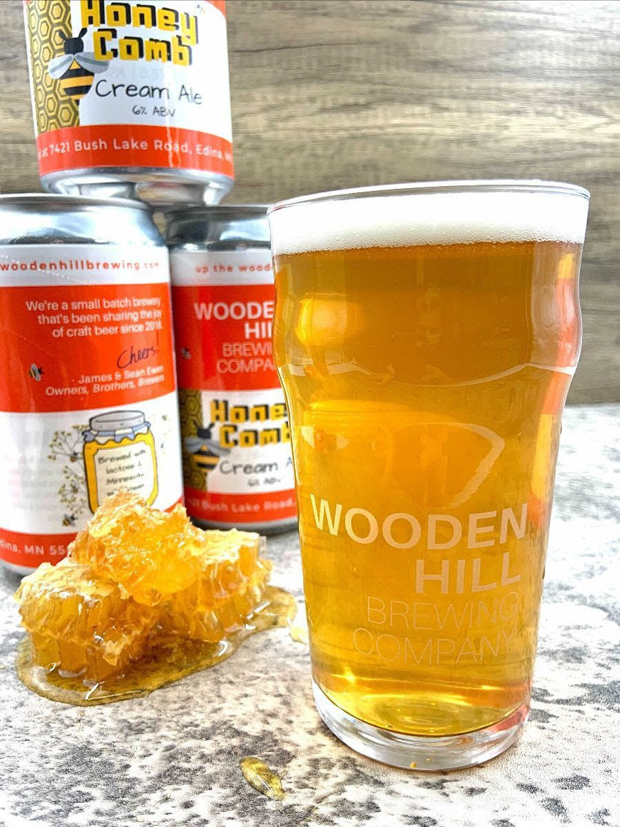 Image of a pint of light beer with cans of beer and a pile of real honeycomb in the background.