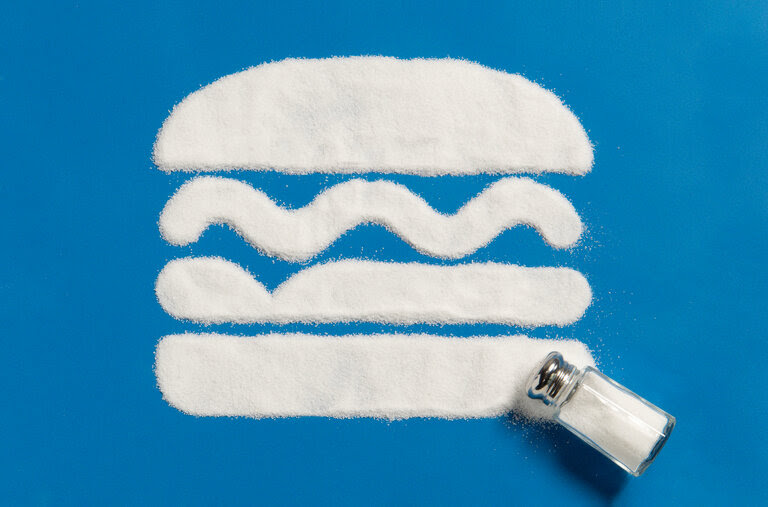 An illustration of salt in the shape of a cheeseburger on a blue background with a tipped over saltshaker on the bottom right. 