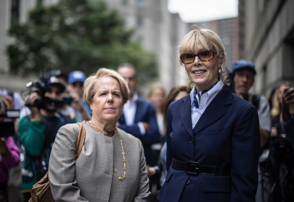 E. Jean Carroll is pictured wearing a blue blazer and sunglasses. She stand next to another woman who is wearing a gray jacket.