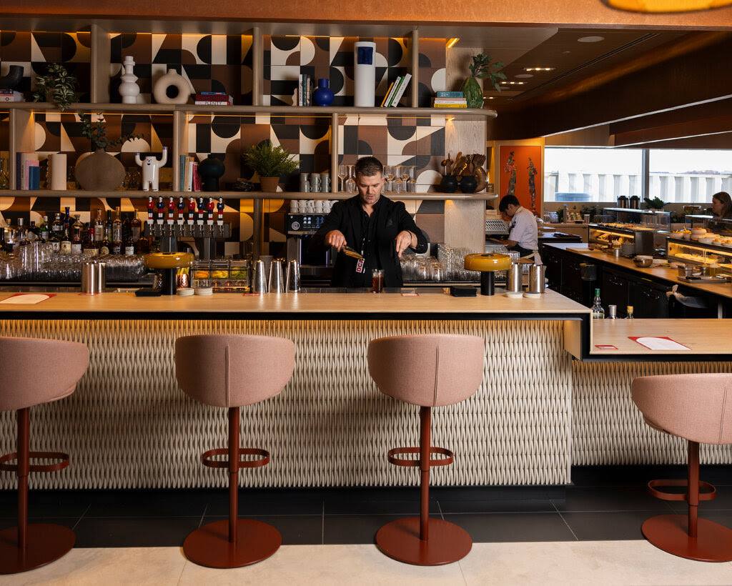 A bartender wearing a black T-shirt prepares a cocktail behind a bar. In front of the bar are pink-and-brown bar seats.