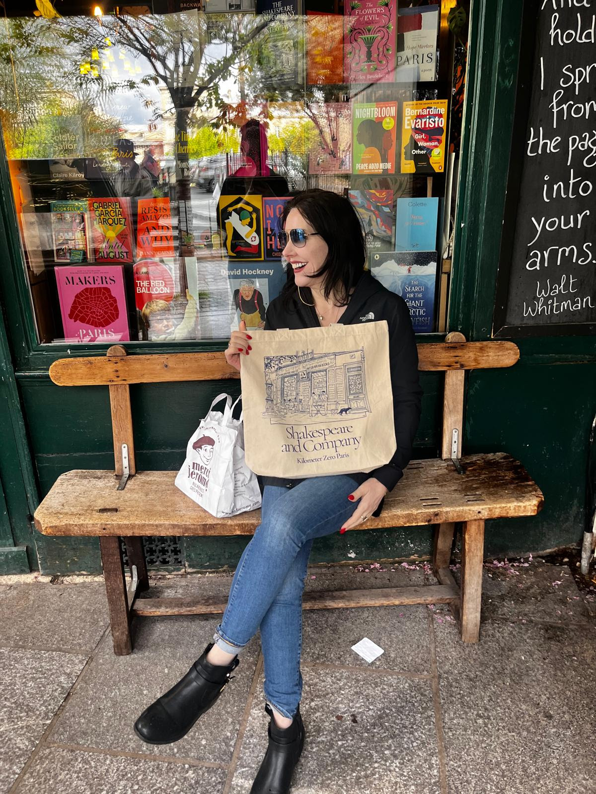 Mary Moynihan sits on a bench outside the Shakespeare and Company independent bookstore in Paris. She faces to her right, laughing.