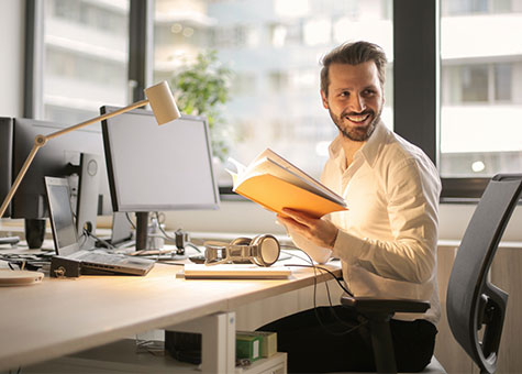 The photo depicts a cheerful man in a modern office, surrounded by computers and office supplies, suggesting a profession in digital technology or data analysis.