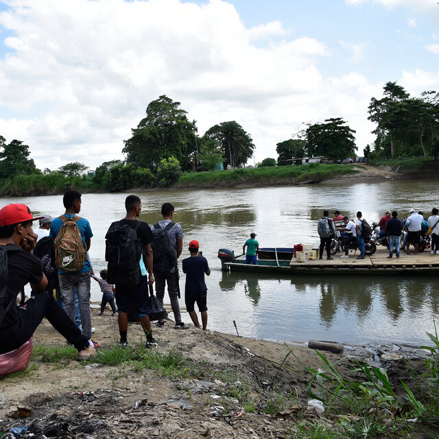 Refugiados en una barca cruzando un río mientras otros esperan en la orilla.