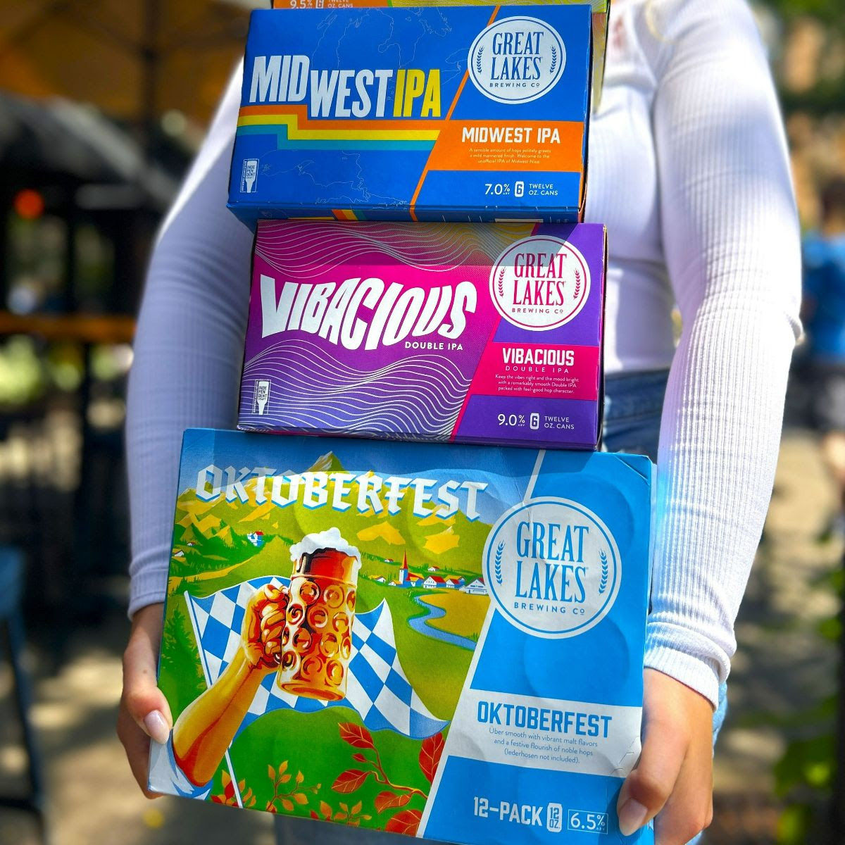 Woman holding 12 oz. Can 12-Pack of Oktoberfest, and 6-Packs of Vibacious Double IPA and Midwest IPA.