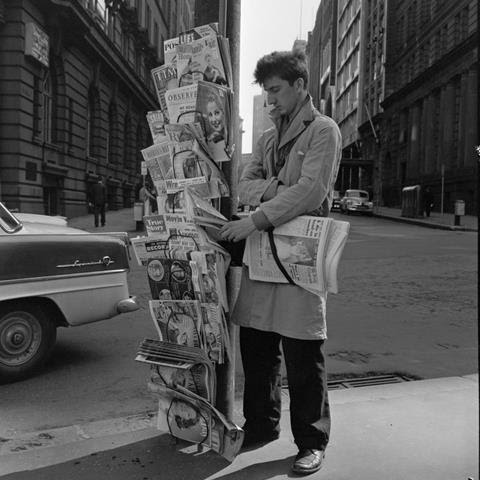 Black and white photo of a man standing at a newsstand on a street.