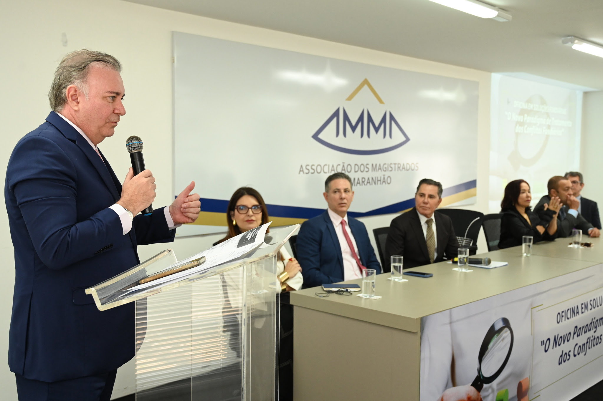 A imagem mostra uma conferência ou reunião formal com várias pessoas sentadas em cadeiras, assistindo a uma apresentação. Na frente da sala, há uma mesa com seis pessoas sentadas, e uma pessoa em pé ao lado de um púlpito, aparentemente fazendo uma apresentação ou discurso. No fundo, há um banner com o logotipo e o nome "ASSOCIAÇÃO DOS MAGISTRADOS DO MARANHÃO". Abaixo do banner, há uma faixa com o texto "OFICINA EM SOLUÇÕES FUNDIÁRIAS". A sala está bem iluminada e as pessoas parecem estar prestando atenção à apresentação.