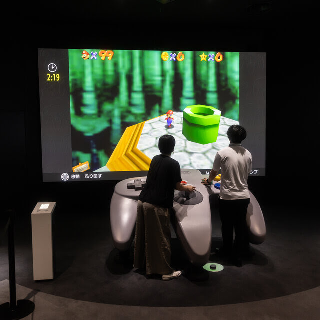 Two people operate a giant Nintendo 64 controller while playing a Mario video game.