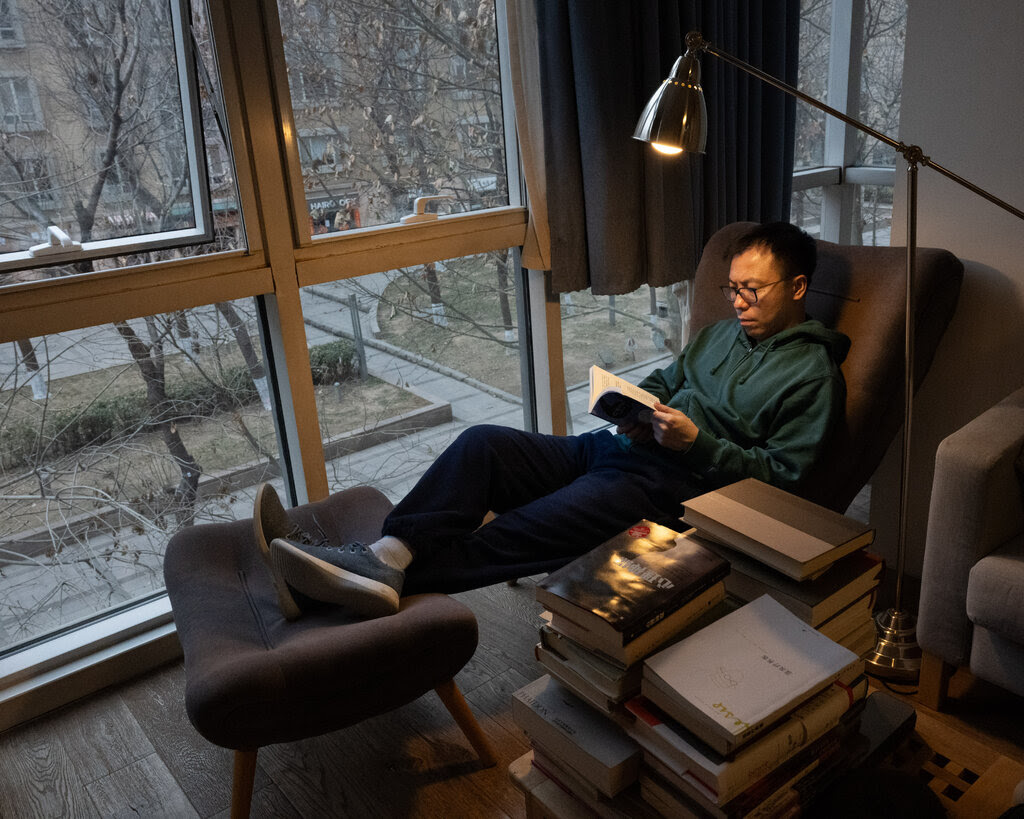 A bespectacled Shuang Xuetao reading in a comfortable chair by large windows. He’s reclining, with his feet, clad in sneakers, resting on a footstool. 