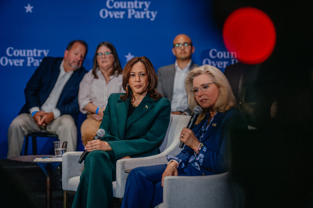 Kamala Harris, in a green suit, and Liz Cheney, in a blue outfit, hold microphones while sitting on a stage.