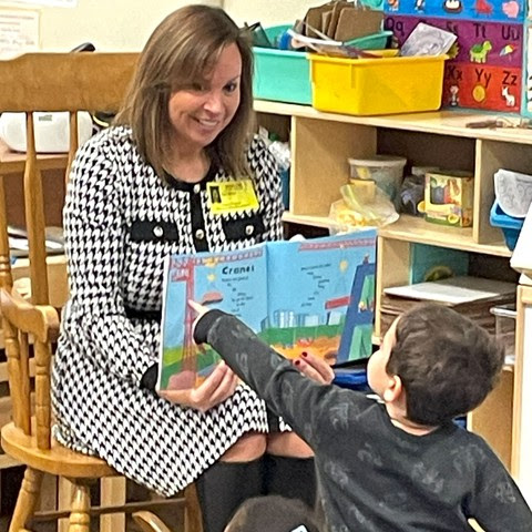 a photo of Chancellor Miller sitting in a rocking chair with the storybook open, a young boy points to the picture