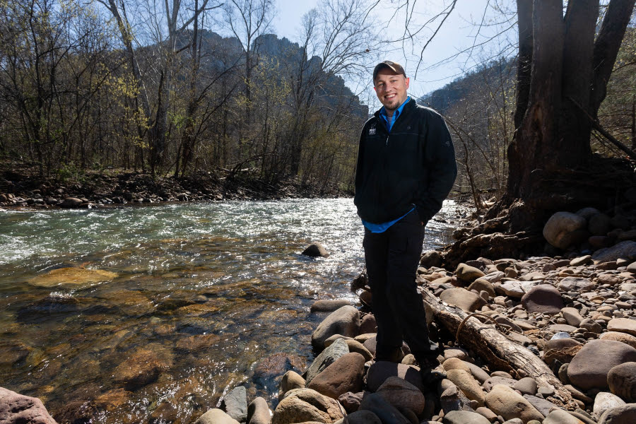 Dustin is a white man wearing a hat and a black jacket. He stands in front of a river during late fall.