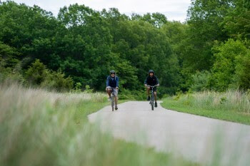 Two people cycle down a paved trail in summer.